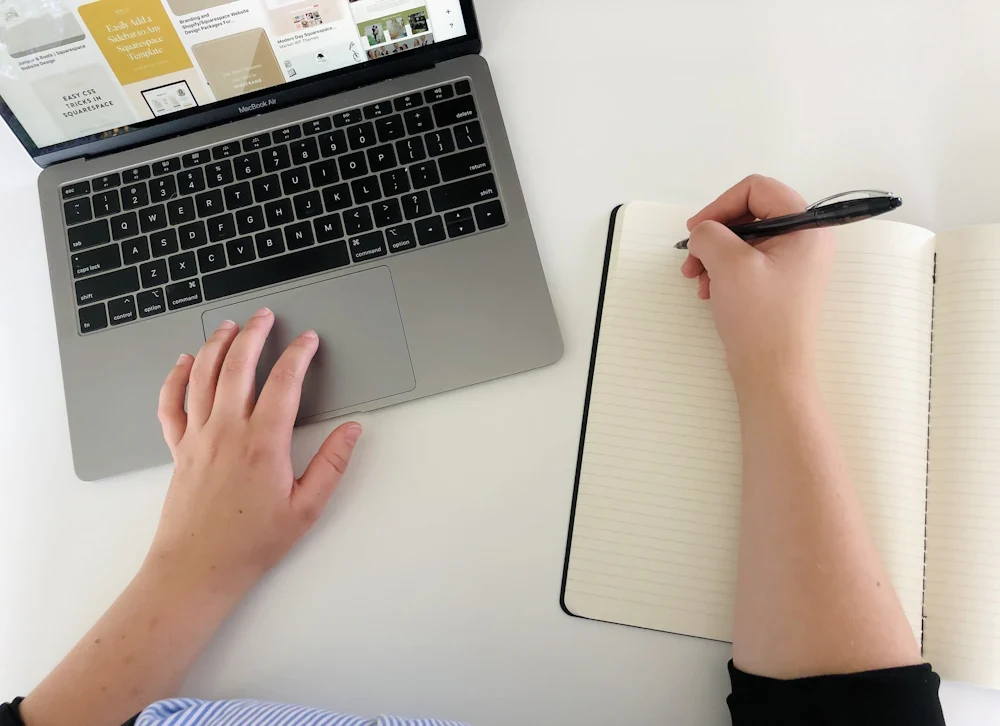 Hands taking notes beside a laptop; illustrative stock photograph by MJ Duford / Unsplash.